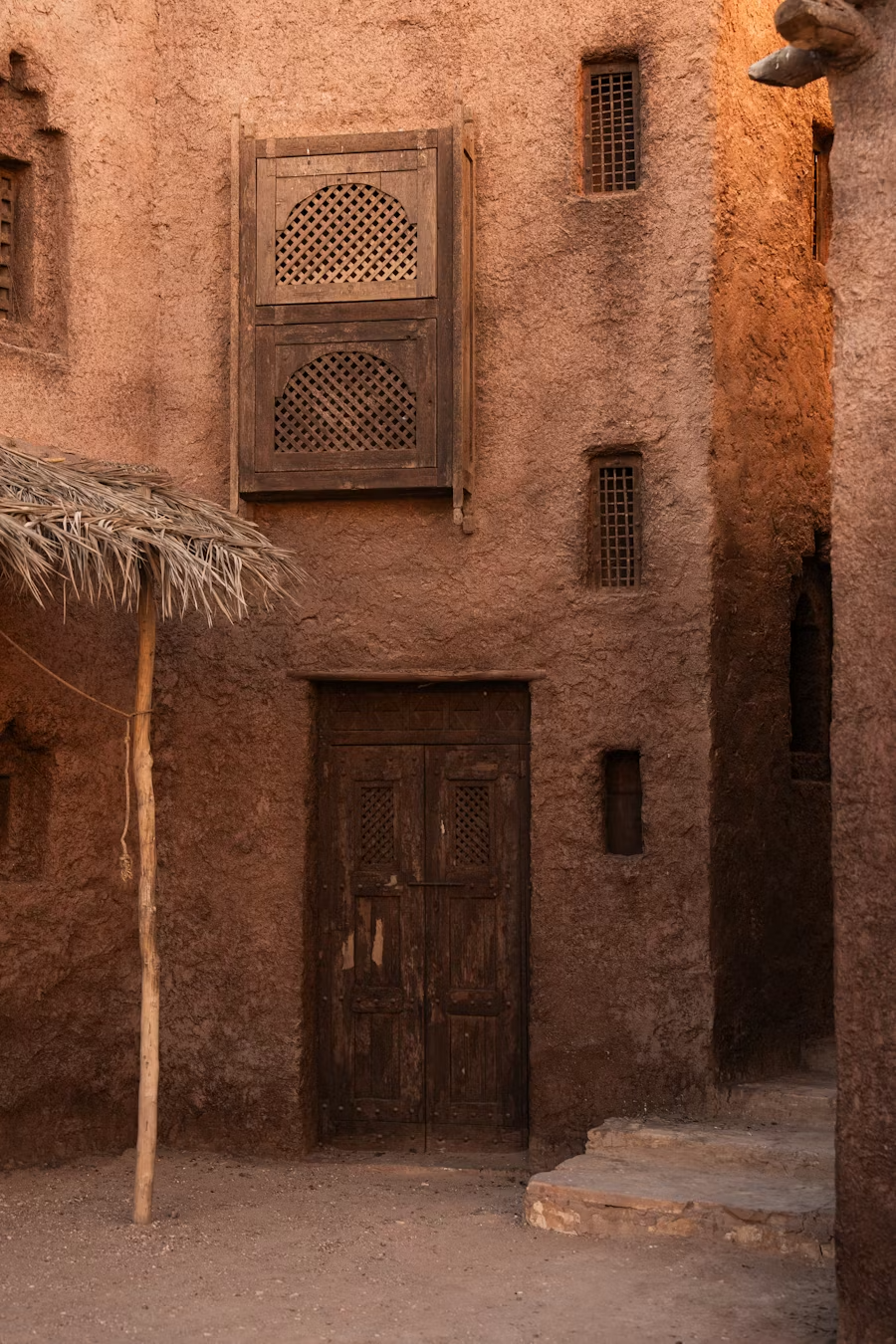 An old rustic building with a wooden door, small rectangular windows with grills, a thatched umbrella, and pinkish-brown textured walls.