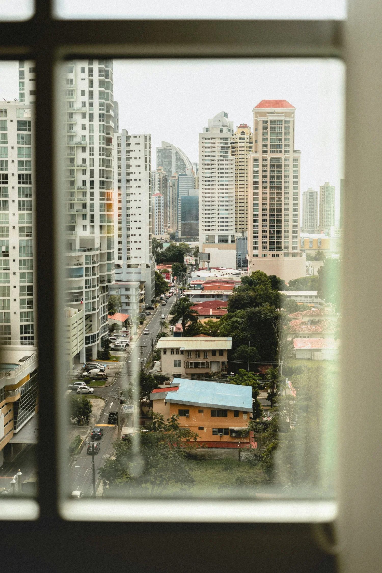 Cityscape viewed through a window with bars, showing tall modern buildings, smaller houses, and trees in an urban area.