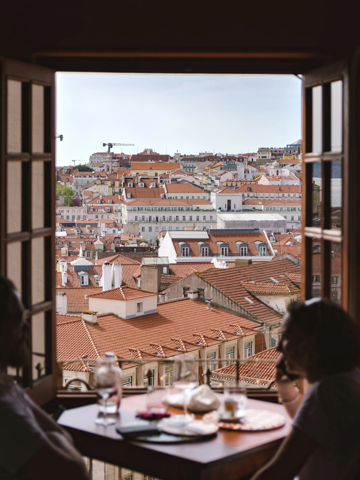 A view of a European city skyline through a window, with red-tiled rooftops and various buildings, as seen from a table inside a room, where two people are dining.