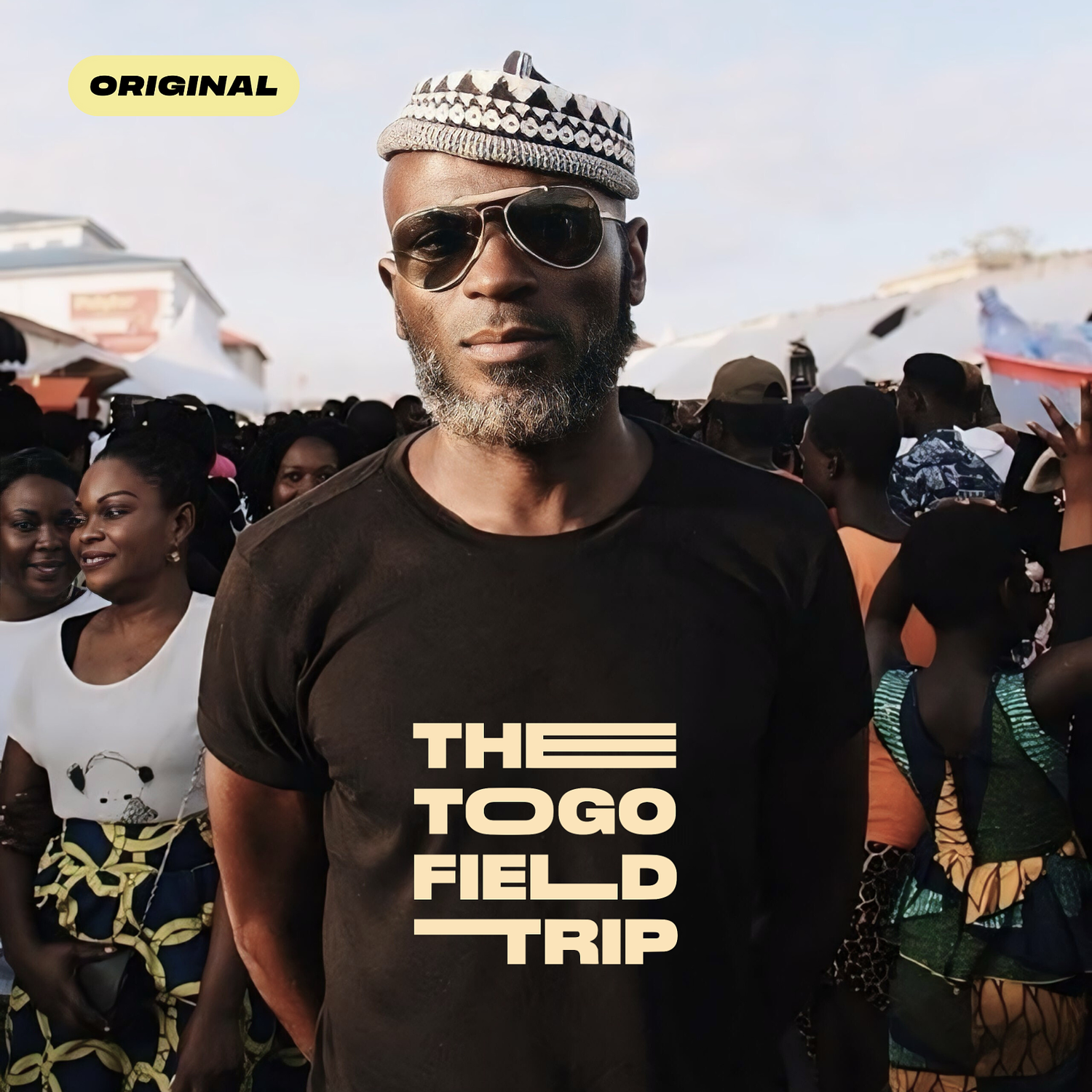 A man with sunglasses and a patterned hat standing among a crowd of women at an outdoor event with white tents in the background. The text on the image reads 'The Togo Field Trip' and 'Original' in the top left corner. Hyperlocal World of Travel.