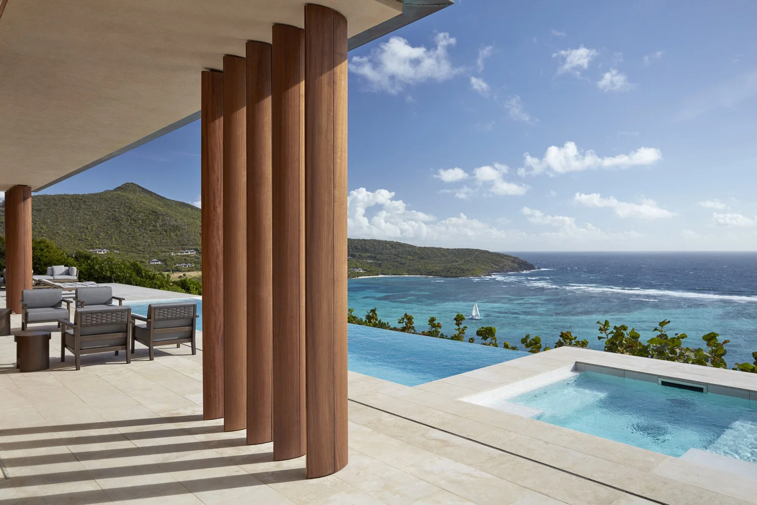 Luxury outdoor patio with wooden columns, seating area, infinity pool, and ocean view with sailboat, green hills, and partly cloudy sky.