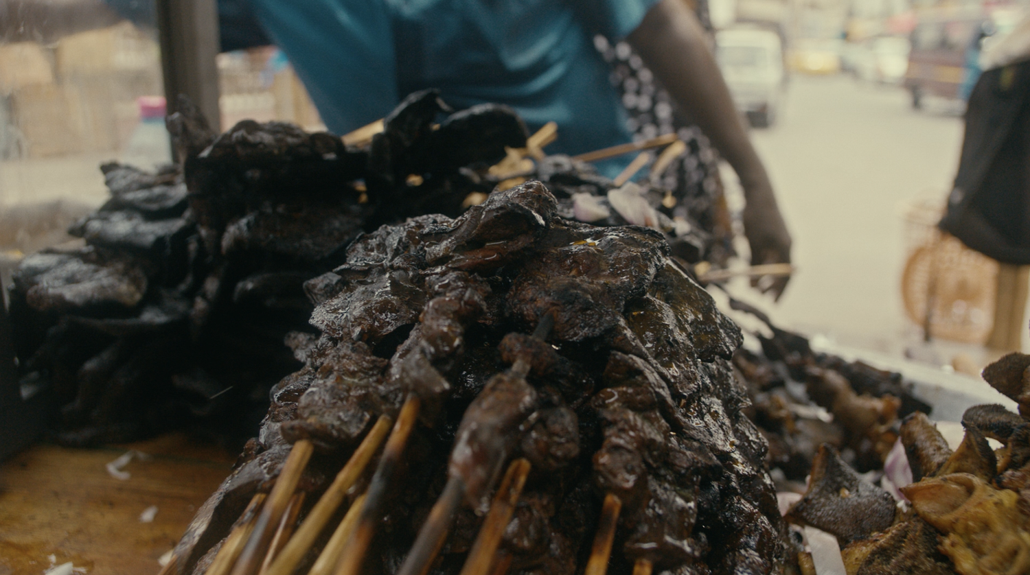 Close-up of skewered grilled meats on display at a street food stall, with a background of a street scene and a person in a blue shirt.