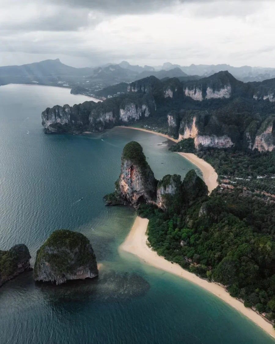 Aerial view of a coastal landscape with a beach, cliffs, and green forested areas, under cloudy skies.