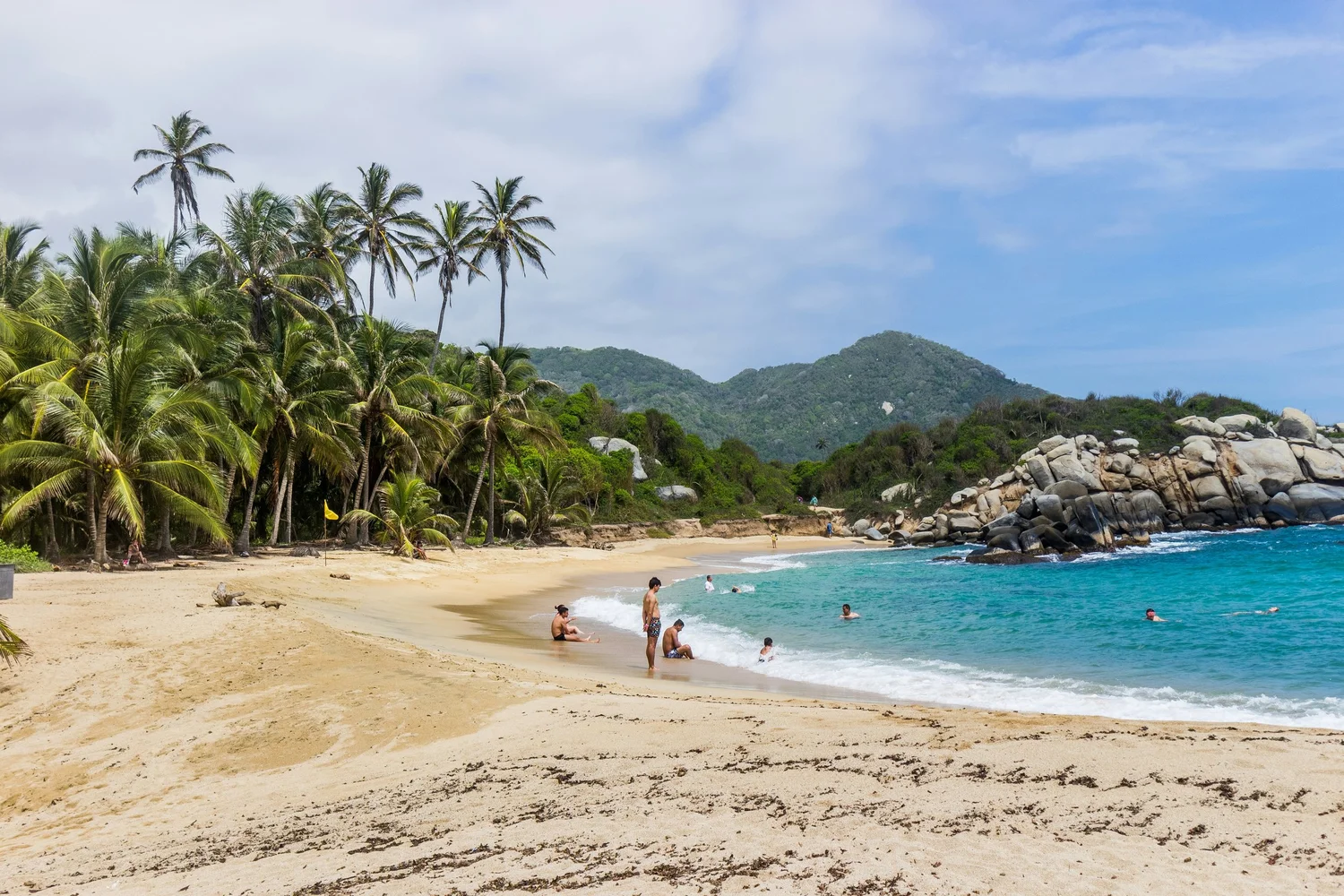 Beach with palm trees, rocks, and people swimming and relaxing by the water near lush green hills and mountains in the background.