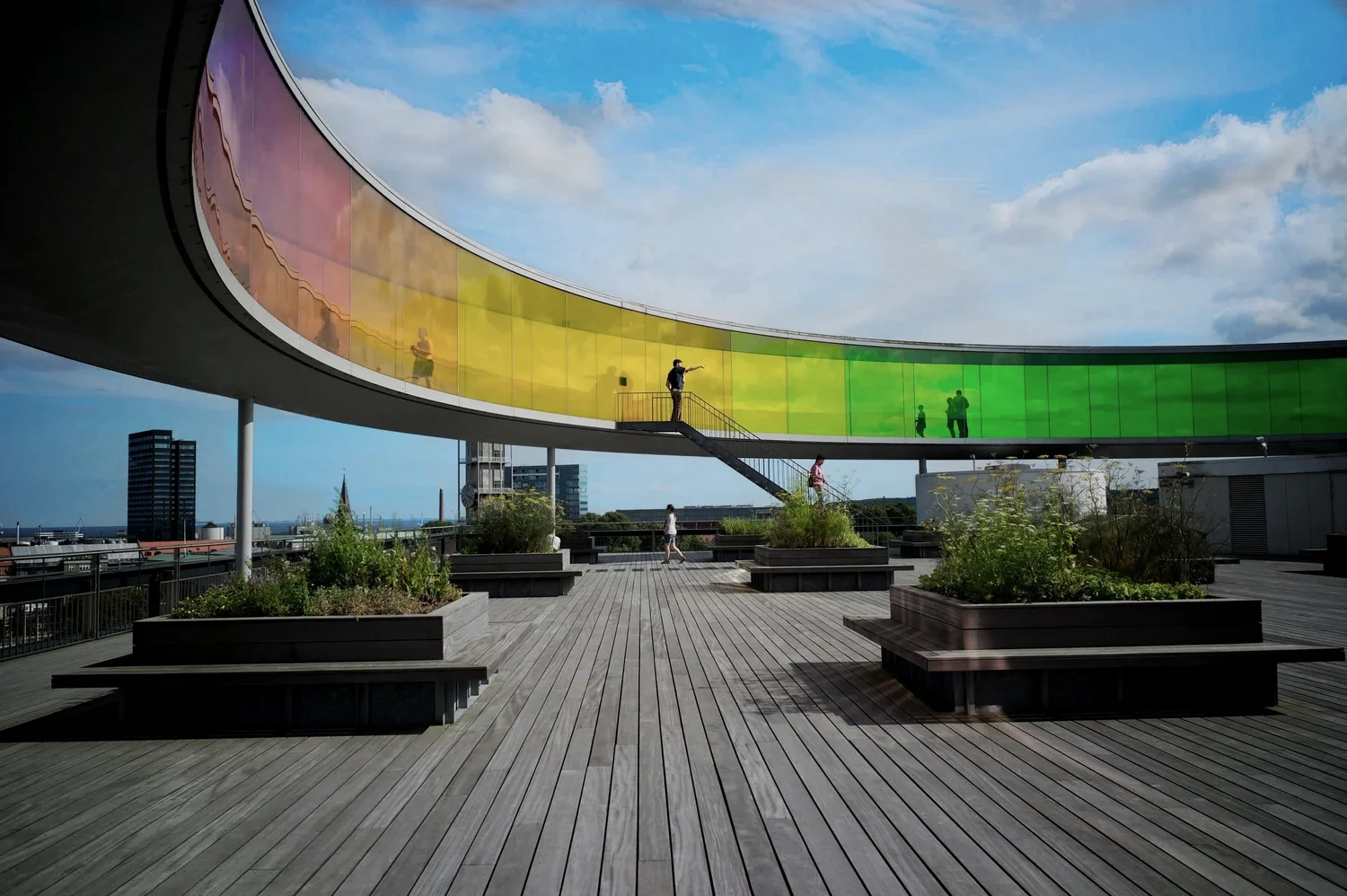 People walking on a rooftop terrace with wooden decking and planter boxes, overlooking city buildings, with a large curved glass structure reflecting the sky, which is partly cloudy.