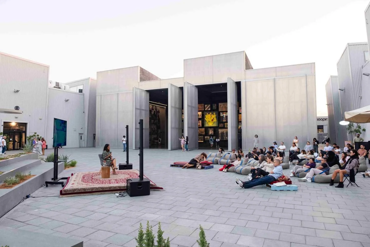 Outdoor art event with a woman performing on a rug with speakers and large screens, audience seated on bean bags and cushions outside modern gallery buildings