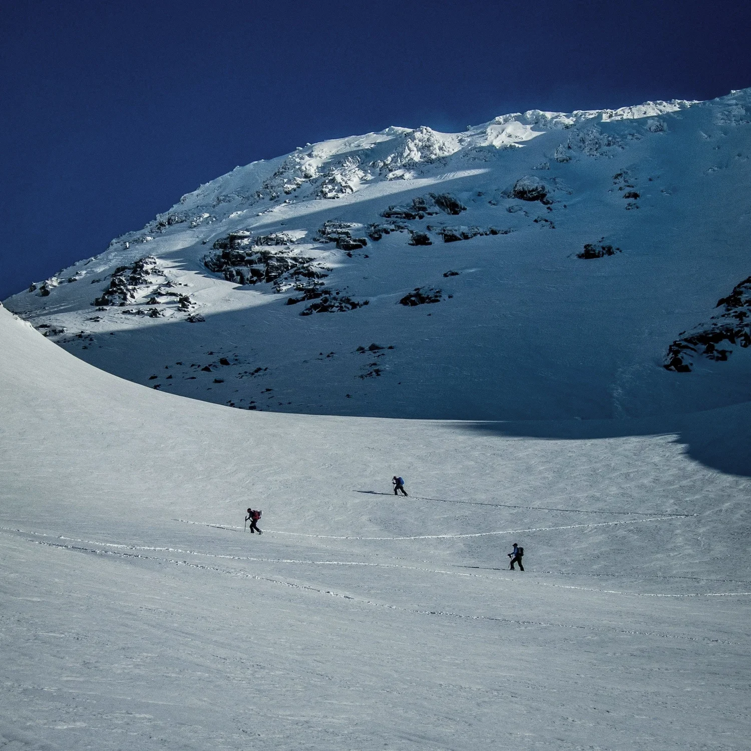 Three people skiing on a snow-covered mountain slope with peaks and rocky outcroppings in the background under a clear blue sky.