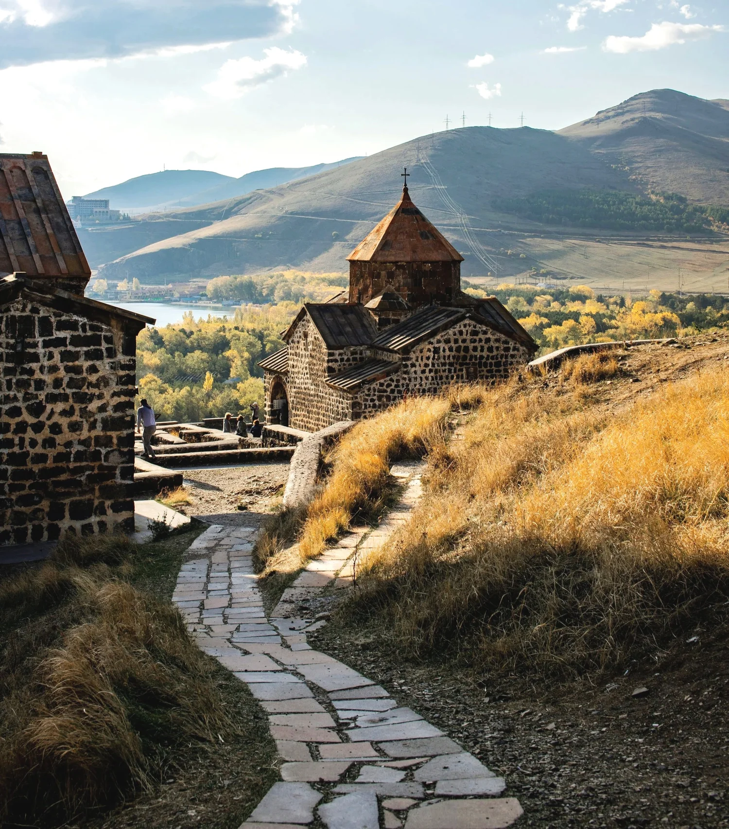 A stone pathway leading up to an old stone church with a metal roof and cross on top, set in a hilly landscape with dry grass, in the background hills and mountains under a partly cloudy sky.