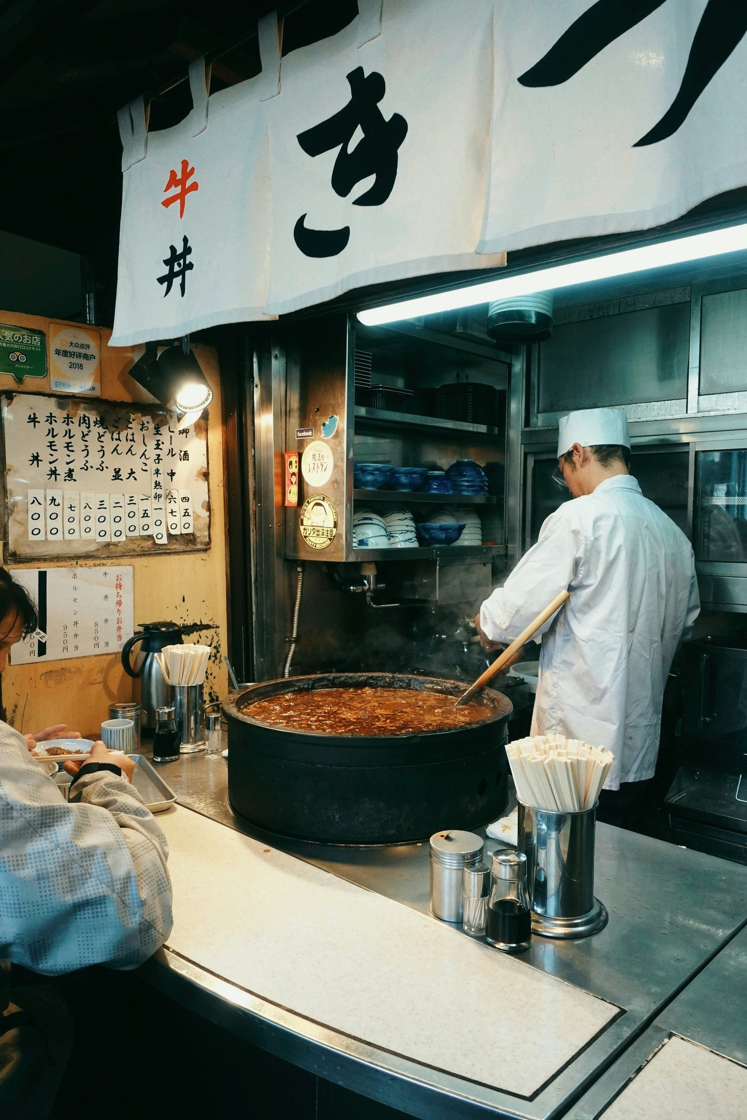 A restaurant kitchen scene with a chef stirring a large pot of stew or sauce, a menu written in Japanese on the wall, and a person sitting at a counter eating.