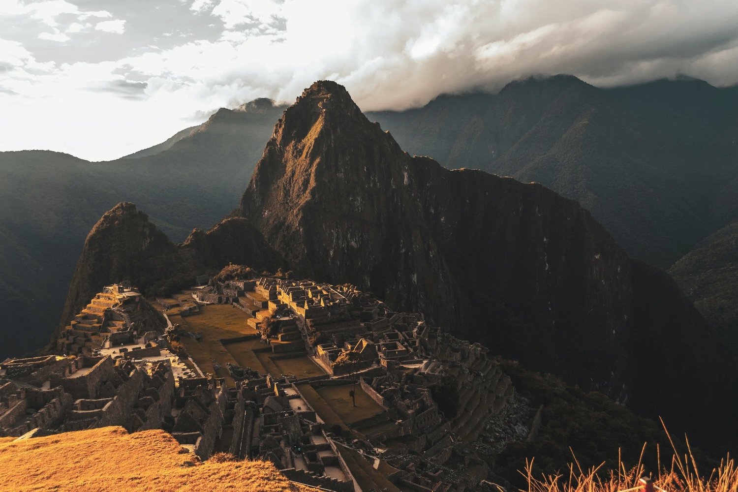 Sunlit view of Machu Picchu with ancient ruins and terraced fields at the base of a steep mountain, with cloudy sky in the background.