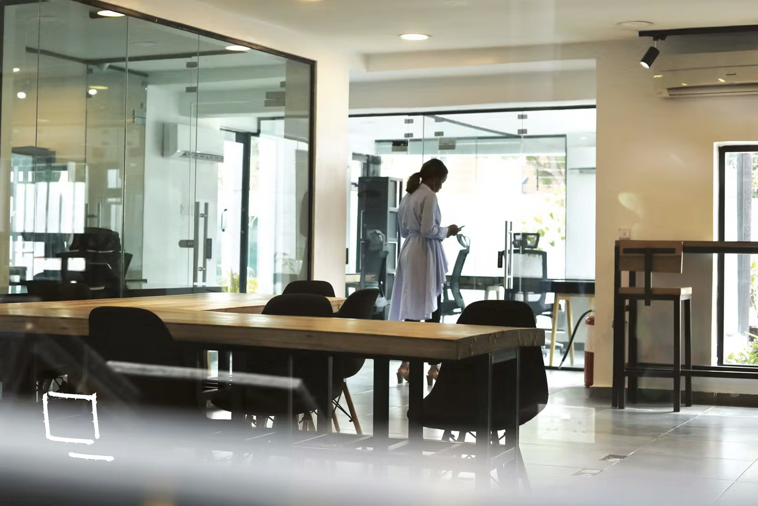 A woman in a white coat standing inside a modern, brightly-lit office or conference room, looking at her phone near a glass wall and table.