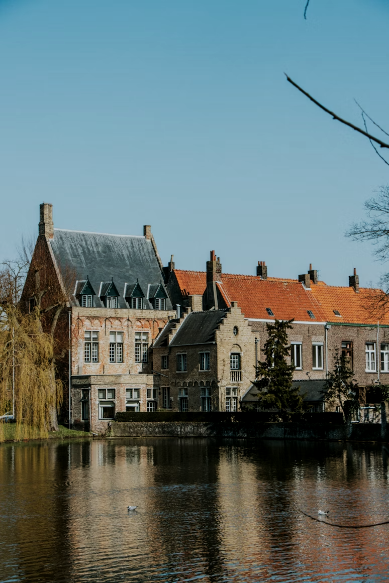 Historic European-style buildings beside water with reflections, some with red-tiled roofs, on clear day with blue sky.