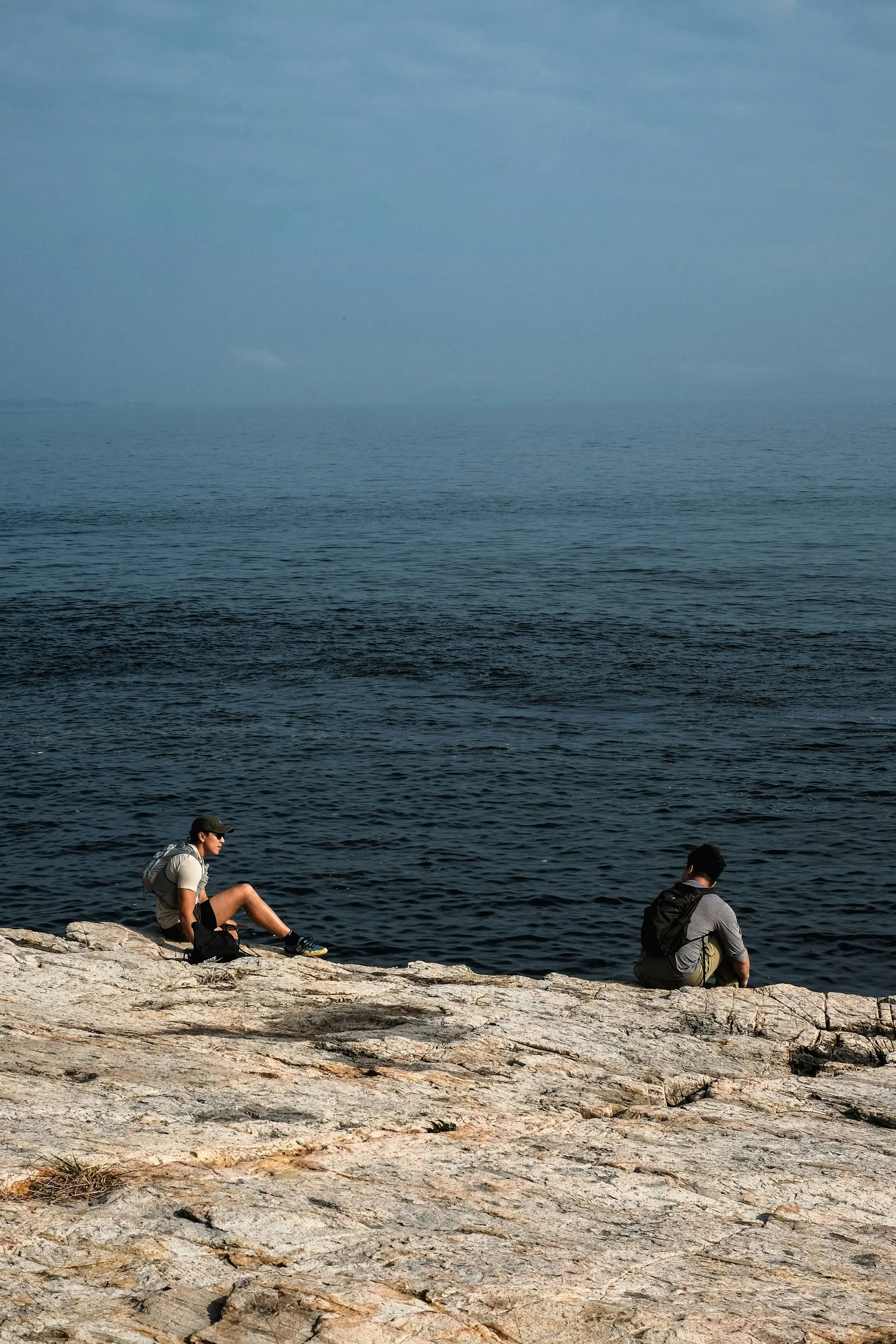 Two people sitting on rocks by the water, fishing in a calm ocean or lake, under a cloudy sky.