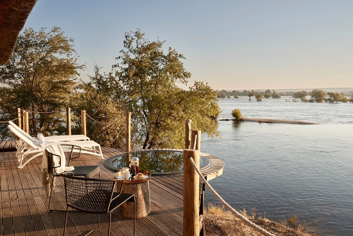 A wooden deck overlooking a river with trees in the background. The deck has lounge chairs, a table with drinks and food, and a rope railing.