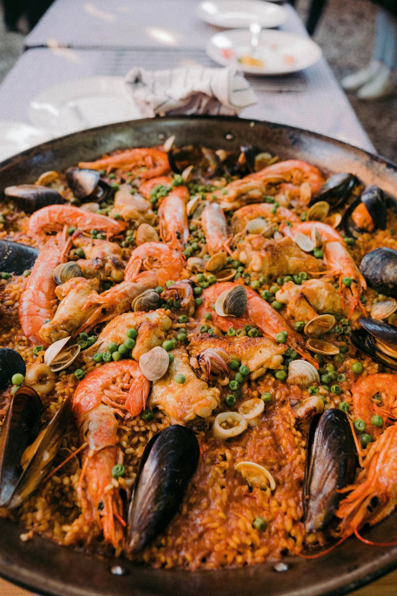 Seafood paella with shrimp, mussels, and green peas on a large skillet, set on a table with plates and utensils in the background.