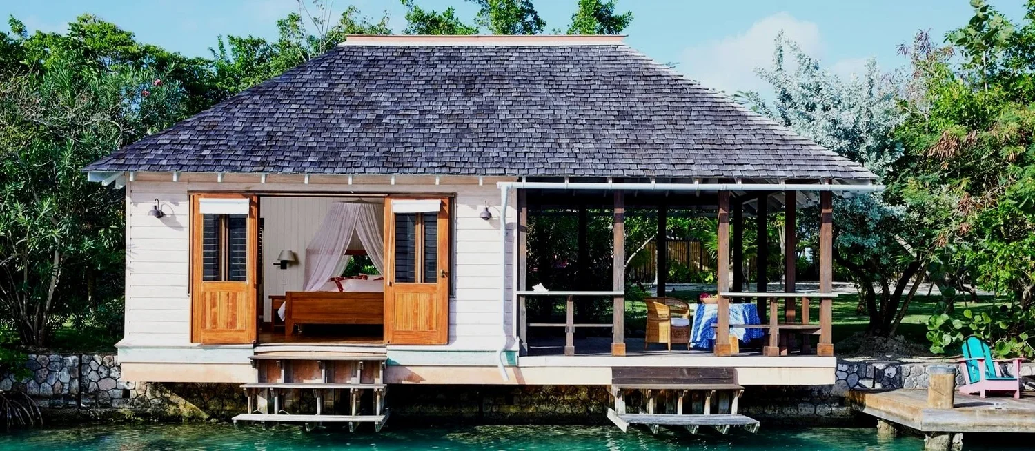 A waterside house with an open sliding door, showing a bedroom inside, and a porch with outdoor furniture, trees, and clear water in the foreground.