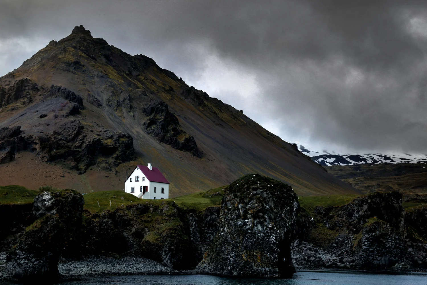 A white house with a red roof sits on a grassy hill by the water's edge, with rocky cliffs and a mountain with patches of snow in the background under a dark, cloudy sky.