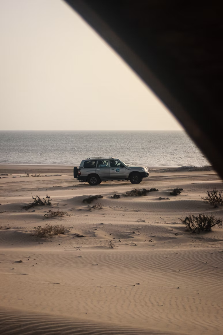 A vehicle parked on a sandy beach near the ocean with some sparse vegetation and a calm cloudy sky.