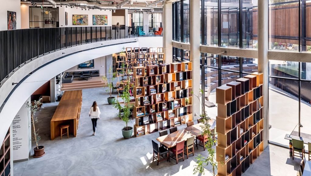 Interior of a modern library or co-working space with large windows, wooden bookshelves, seating areas, and a person walking.