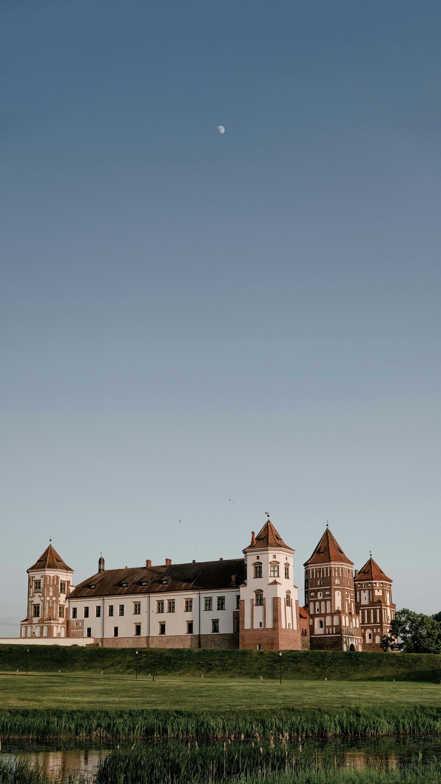 A historic castle with multiple towers and white walls, surrounded by green grass and trees. The sky is clear, with a visible moon and a few birds flying.