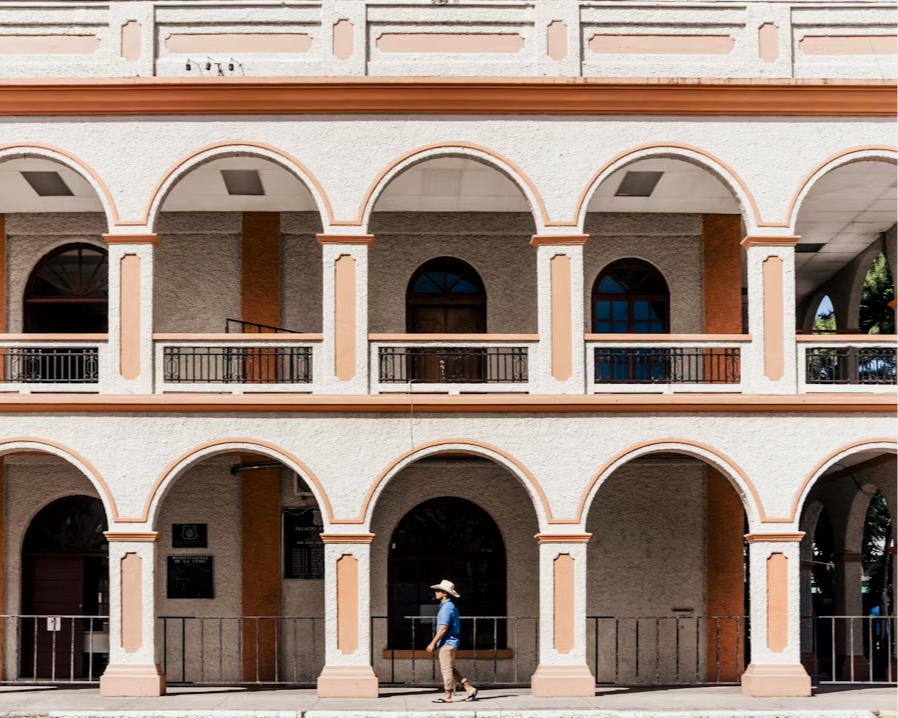 A person walking in front of a building with two floors featuring arches and balconies, with a beige and terracotta color scheme.