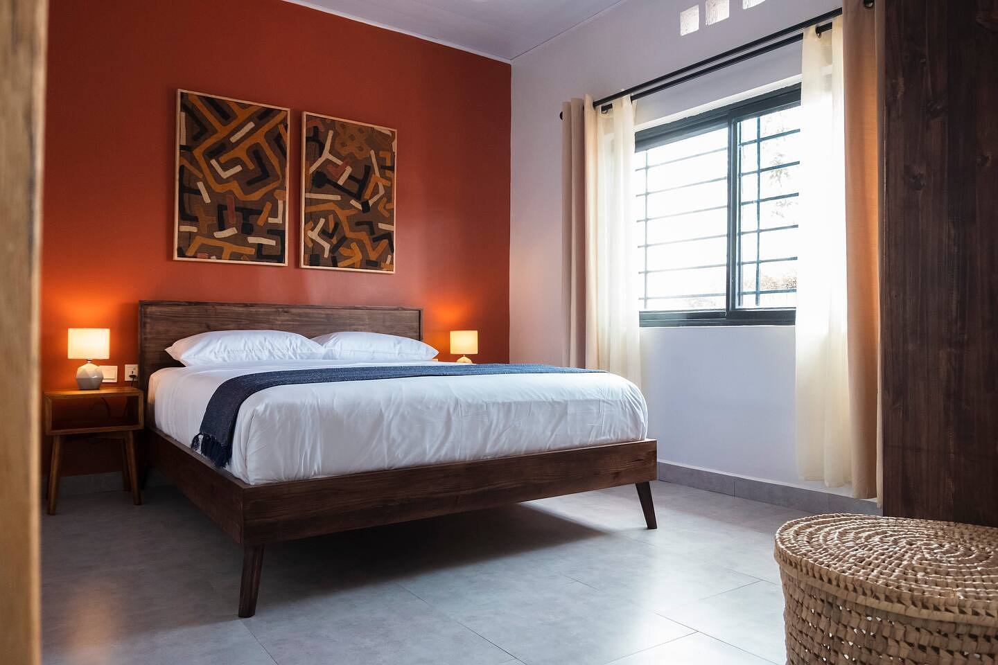 A modern bedroom with a wooden bed, white bedding, two lamps on bedside tables, abstract wall art, a large window with black frame and curtains, and a woven basket in the corner.