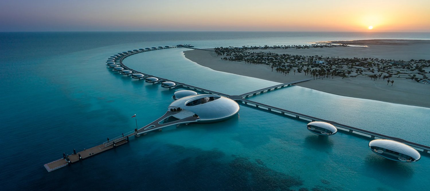 Futuristic coastal resort with sleek, pod-like structures over water connected by bridges, set against a sunset sky with a sandy shoreline and palm trees in the background.