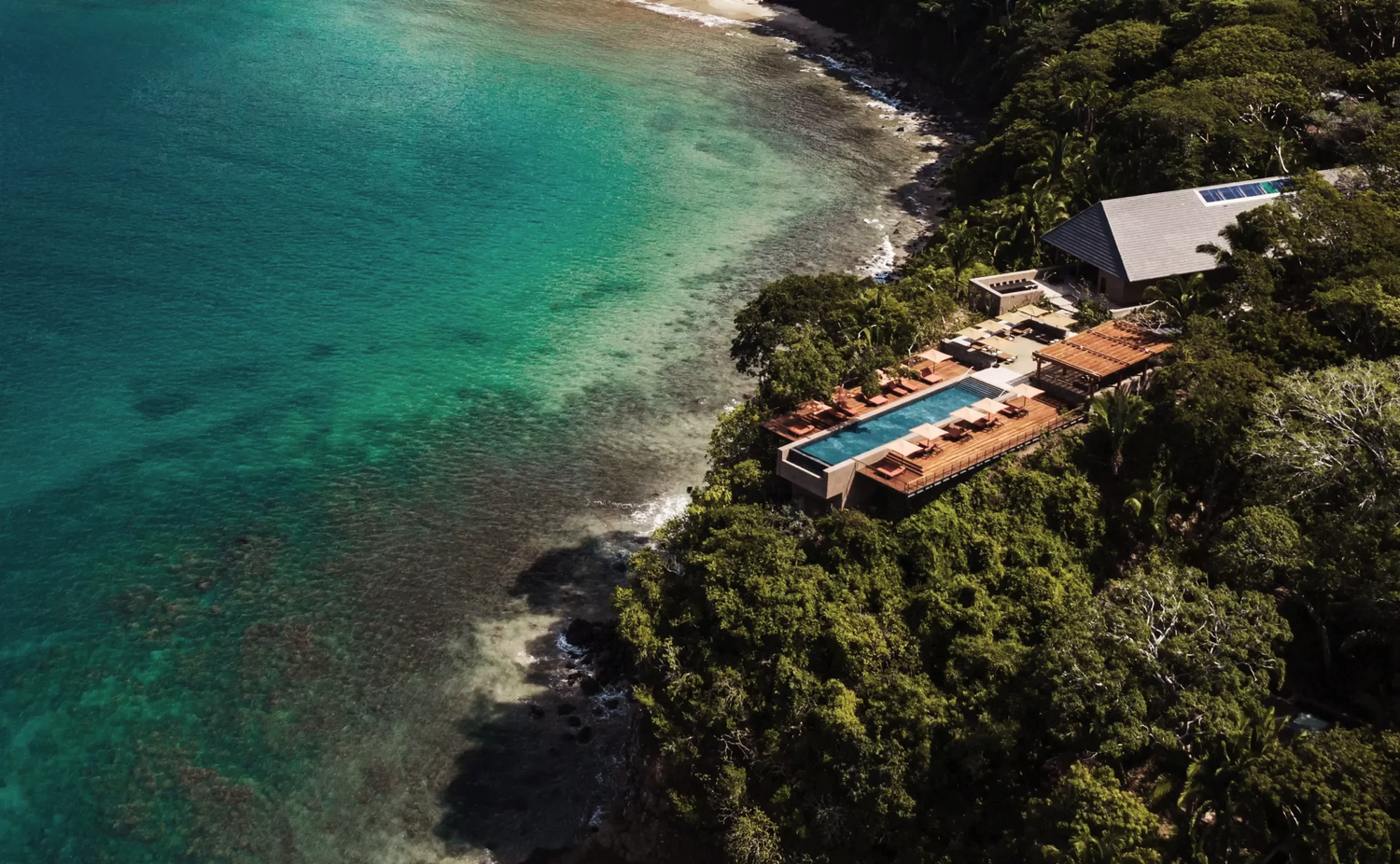 Aerial view of a luxury seaside property with a rooftop pool, surrounded by lush greenery and situated along a clear, turquoise coast.