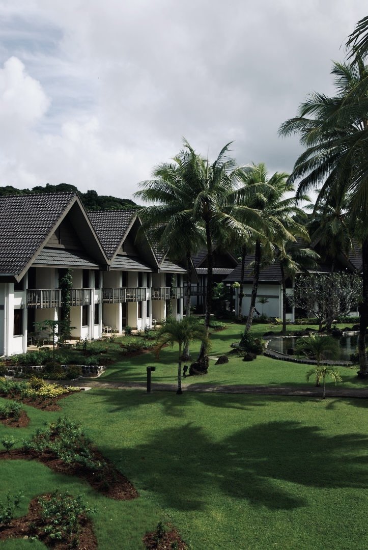 Lush green courtyard with palm trees, garden beds, and a small pond, surrounded by resort-style buildings with balconies and tiled roofs, under a cloudy sky.