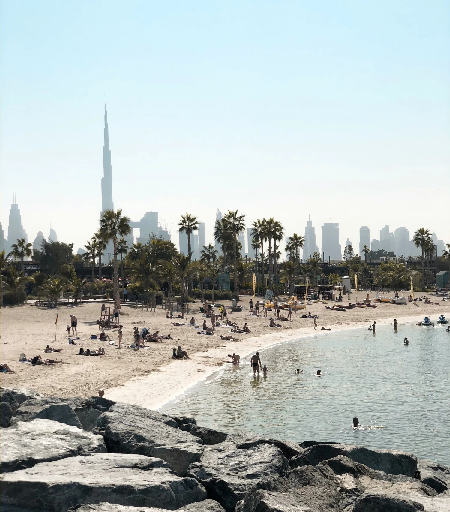 Beach scene in Dubai with people relaxing and swimming, palm trees, and Dubai skyline including the Burj Khalifa in the background.