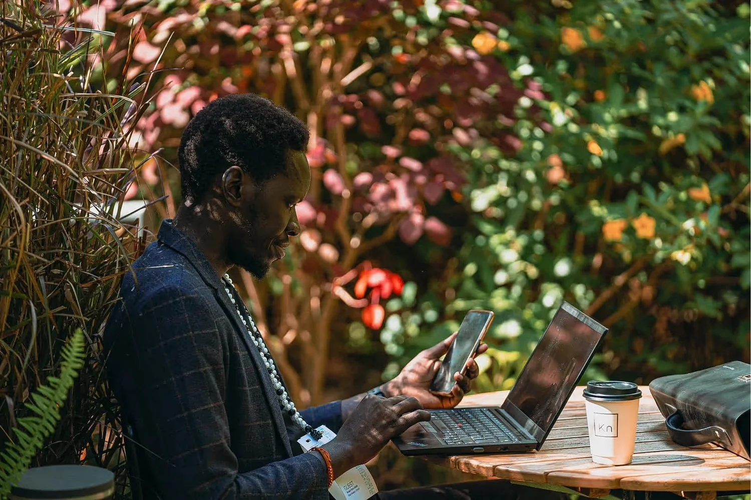 A man with dark skin and curly hair sitting outdoors at a wooden table, using a laptop and a smartphone, with a coffee cup, a bag, and a person’s ID badge nearby, surrounded by lush colorful foliage.