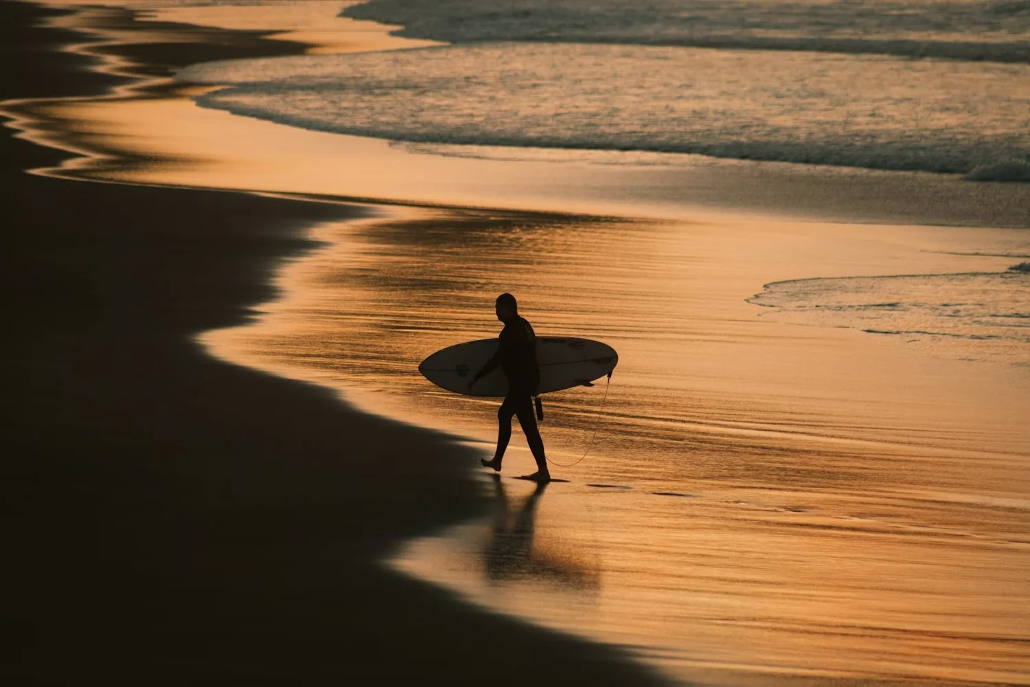 Silhouette of a person carrying a surfboard walking along the shoreline at sunset, with golden reflections on the water.