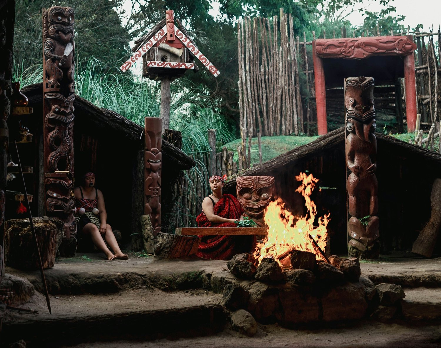 A Maori cultural scene with carved wooden statues, a fire in a circle of rocks, and individuals in traditional attire in an outdoor setting surrounded by greenery.