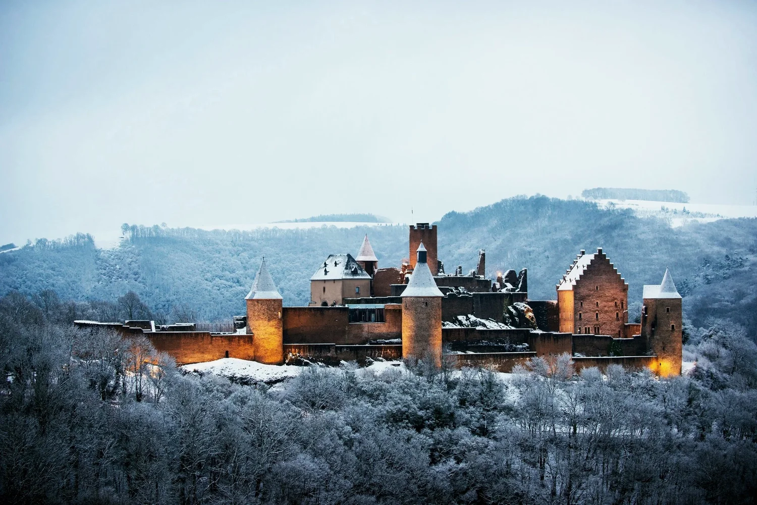 A snowy medieval castle on a hilltop surrounded by snow-covered trees with hills in the background.
