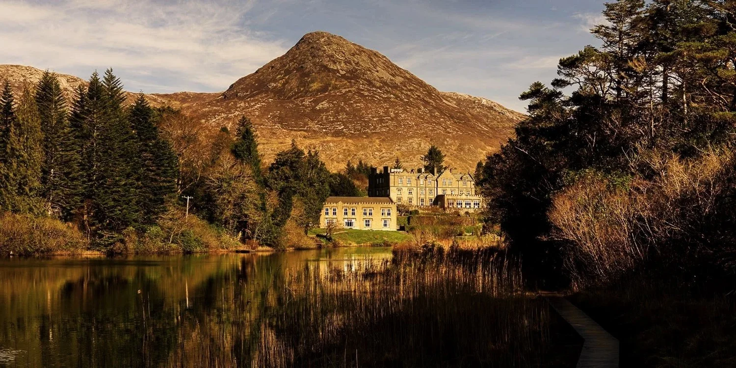 A scenic landscape featuring a large mountain in the background, surrounded by evergreen trees, with a castle-like building in the mid-ground, next to a calm river and a wooden path on the right side.