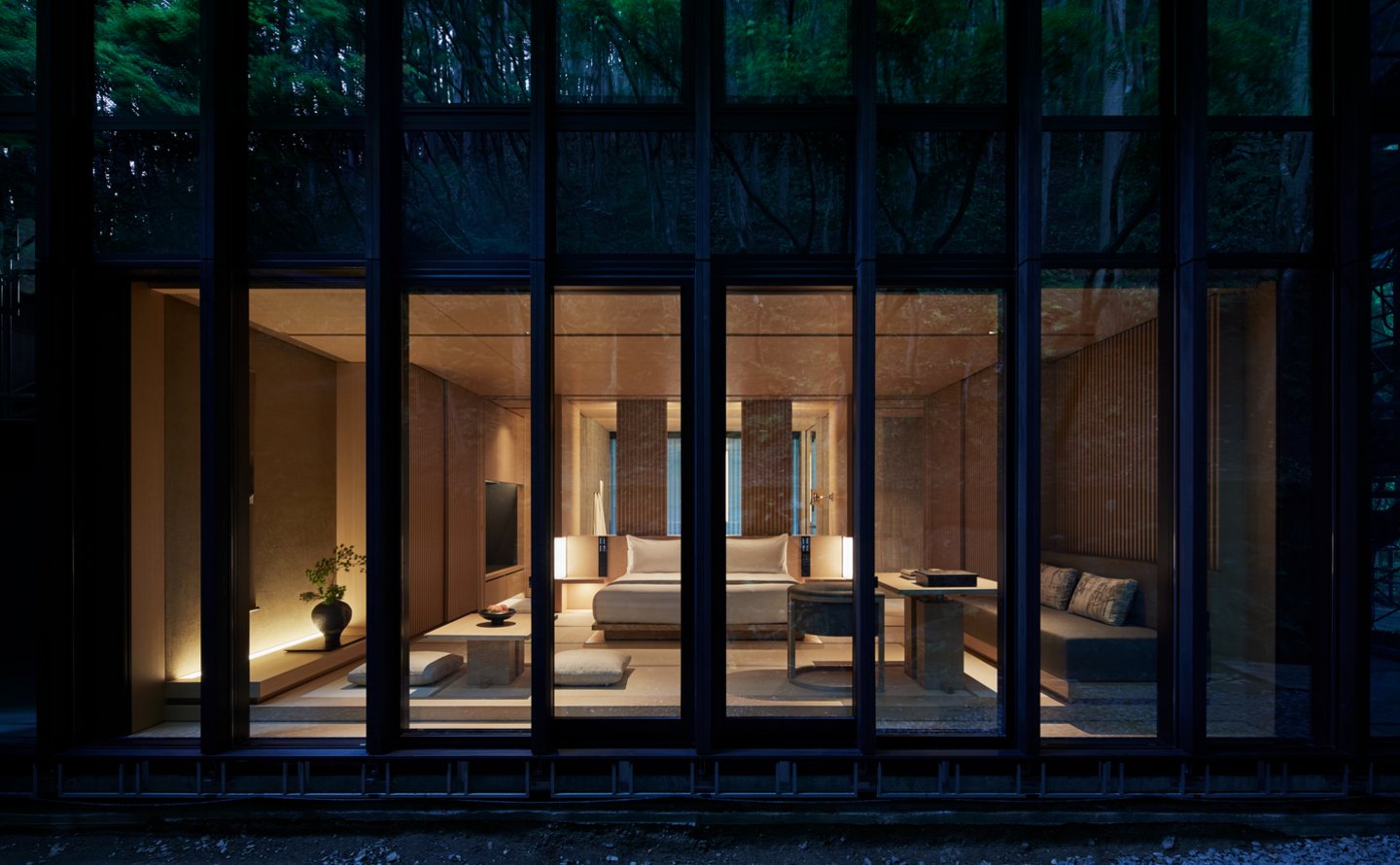 Modern bedroom with warm lighting, large bed, wooden accents, a side table, TV, and a sitting area with cushions, viewed through floor-to-ceiling glass windows at night.