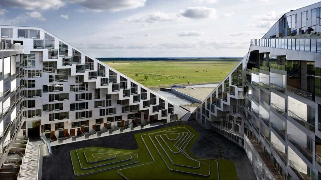 Contemporary residential buildings with geometric designs, surrounding a green maze-like garden, overlooking a flat green field.