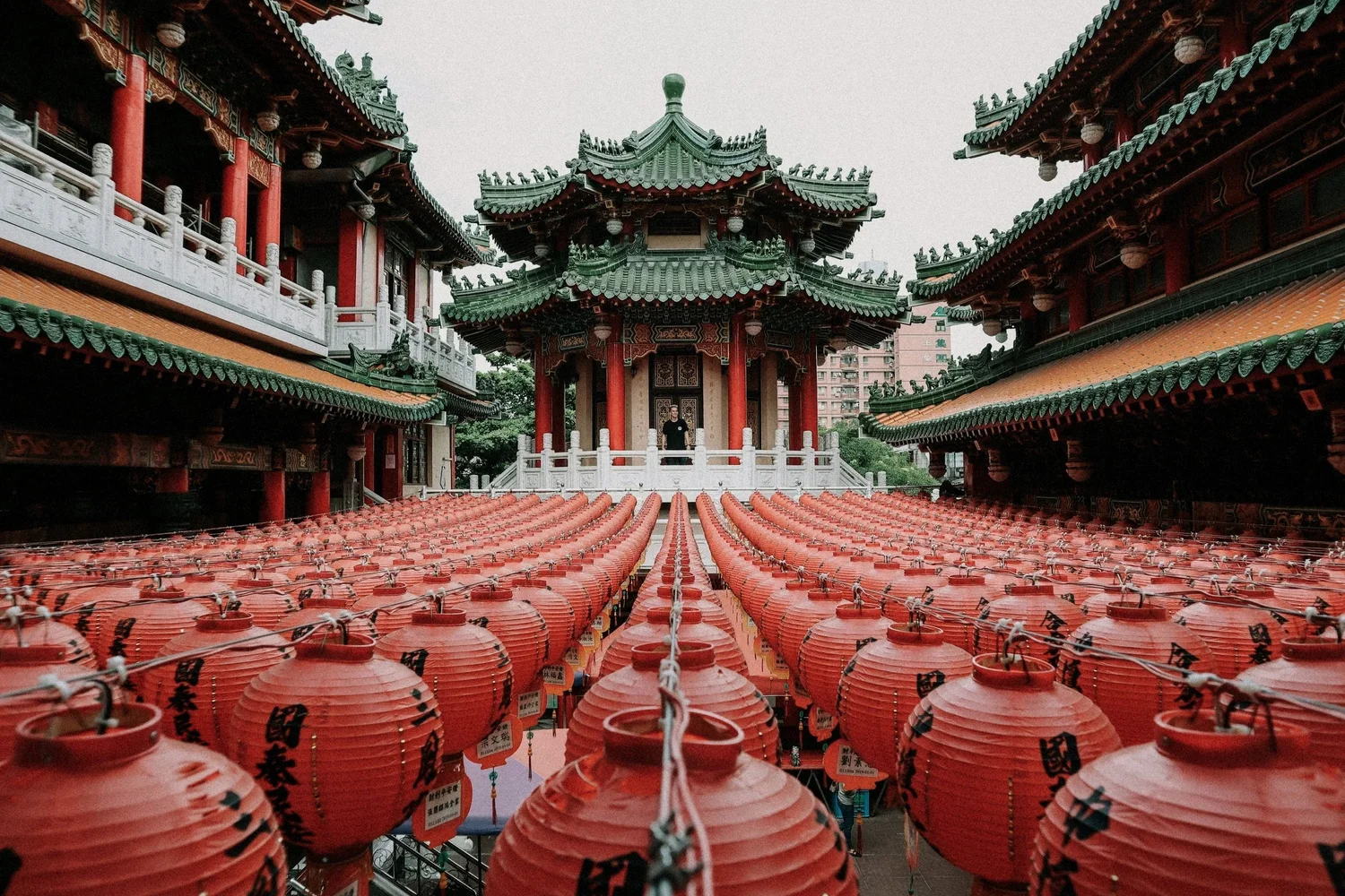 A traditional Asian temple with green tiled roofs and red wooden columns, surrounded by numerous red Chinese lanterns hanging in rows, with a person standing on the temple's balcony.