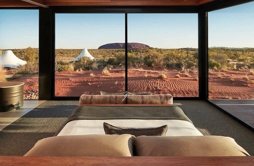 A bedroom with a large window overlooking a desert landscape with sparse vegetation and a rock formation in the distance.