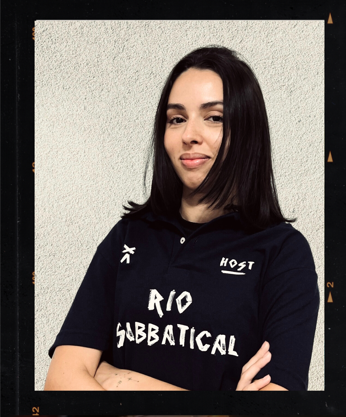 A woman with shoulder-length dark hair wearing a black shirt with white text reading 'RIO SaBBATICAL' and patches, standing against a textured off-white wall, with her arms crossed. Host of Rio Sabbatical by Hyperlocal World of Travel.