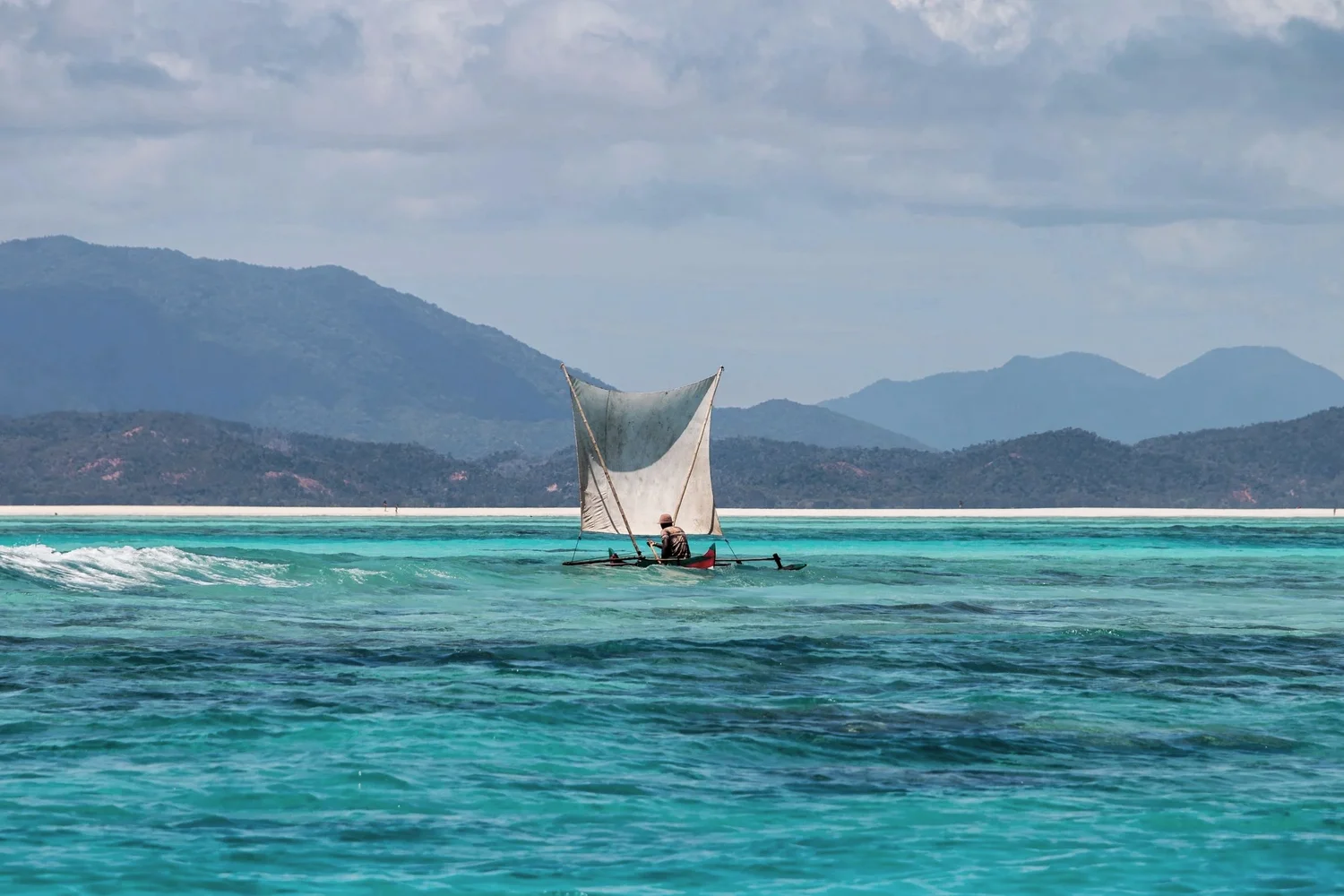 A small sailboat with a single sail is on turquoise water, with mountains and a cloudy sky in the background.