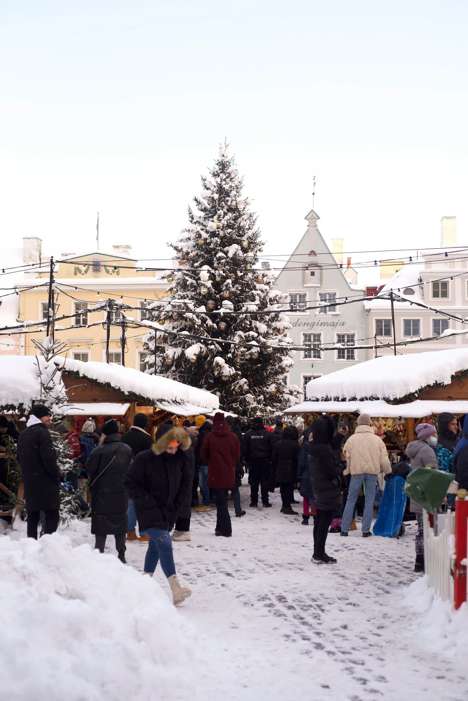 Snow-covered Christmas market with a decorated Christmas tree, wooden stalls, and people bundled up in winter clothing.