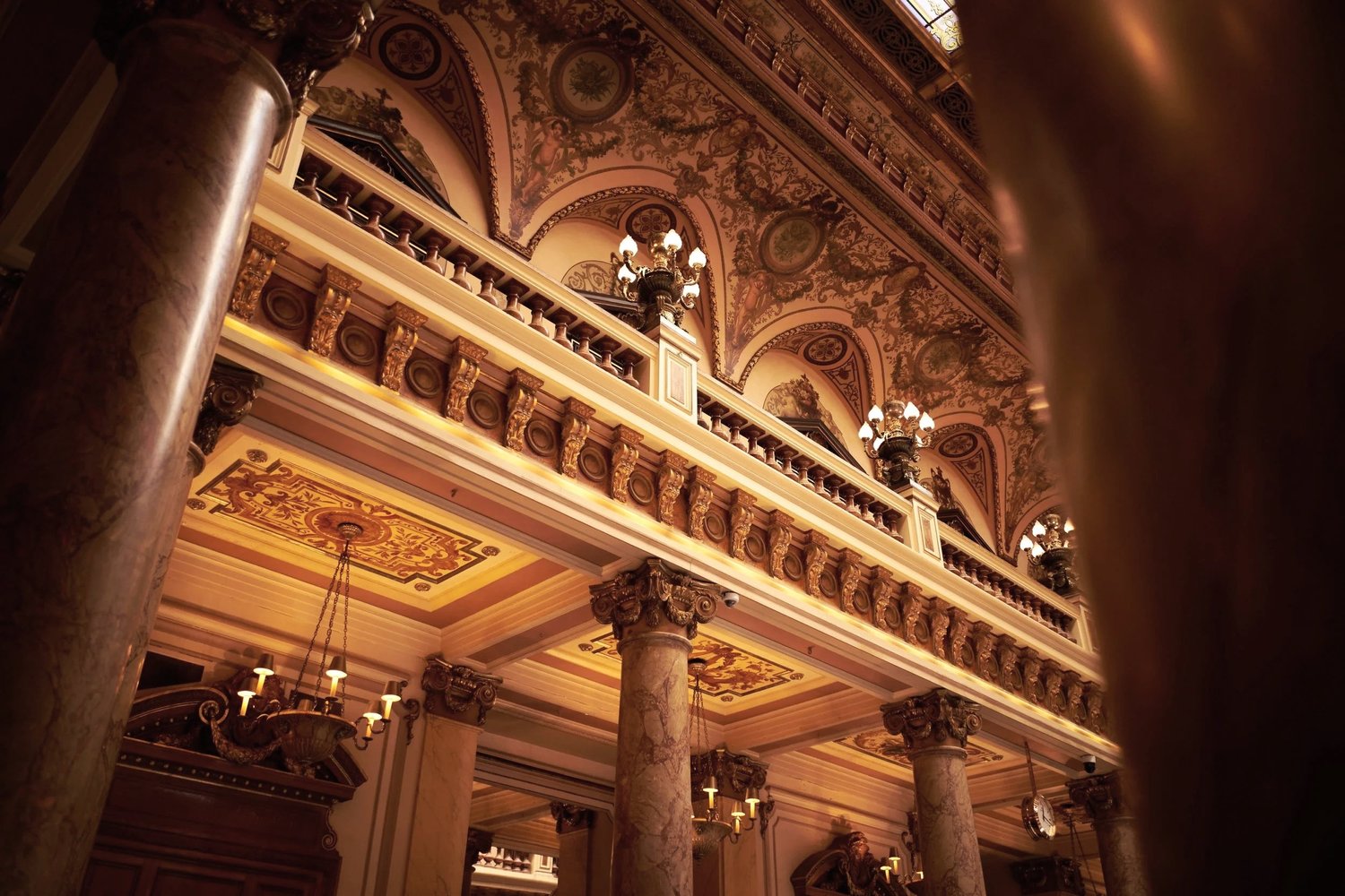 Elegant interior of a historic building featuring ornate columns, intricate ceiling details, chandeliers, and decorative lighting fixtures.