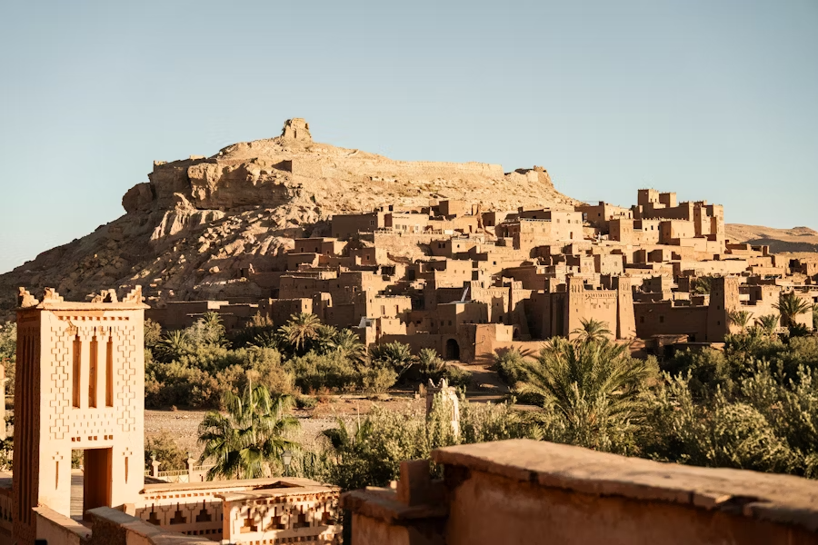 A desert village with traditional mud-brick buildings on a hillside, surrounded by palm trees and greenery.